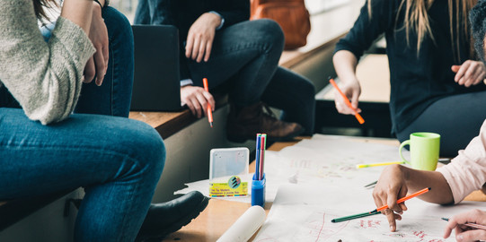 Four people are sitting at a desk writing something on a poster. There are also pencils and a cup on the table.