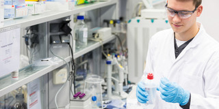 Male trainee in chemistry lab with smock and goggles holds a spray bottle in his hand. 